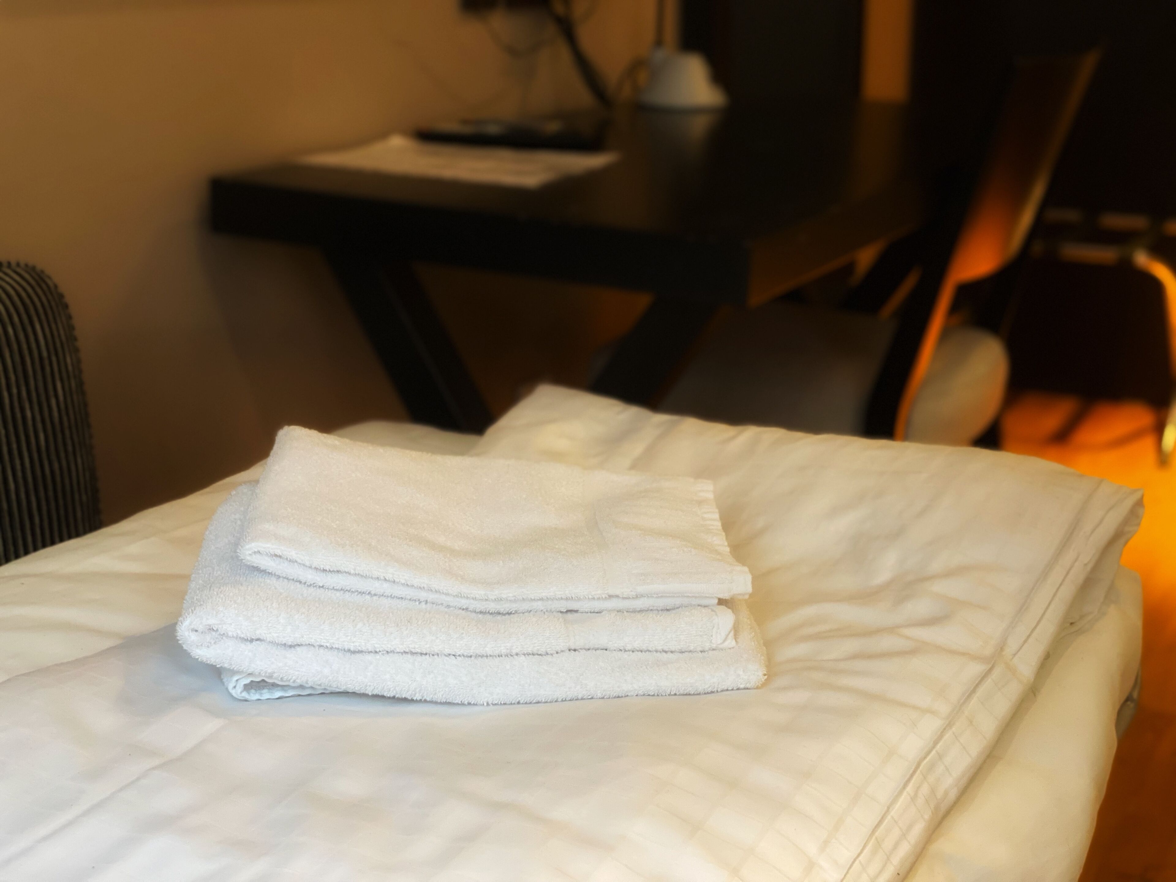 A neatly made bed with folded white towels, a bedside table with a lamp, and a chair in the background.