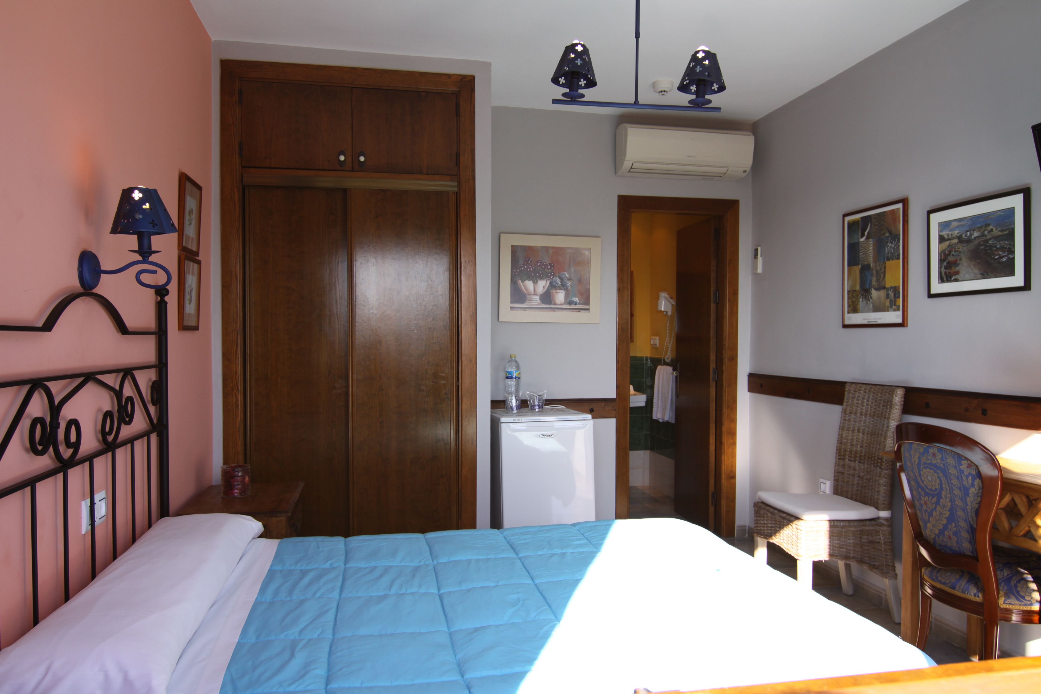 A hotel room with a bed, a desk, a chair, a wardrobe, a refrigerator, and wall-mounted lights.