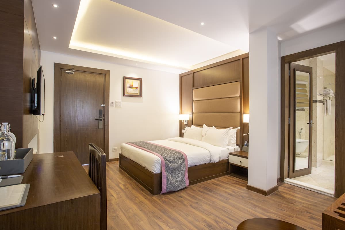 Quarto de hotel com cama, mesa de cabeceira, televisão, chão de madeira e banheiro com chuveiro e vaso sanitário.