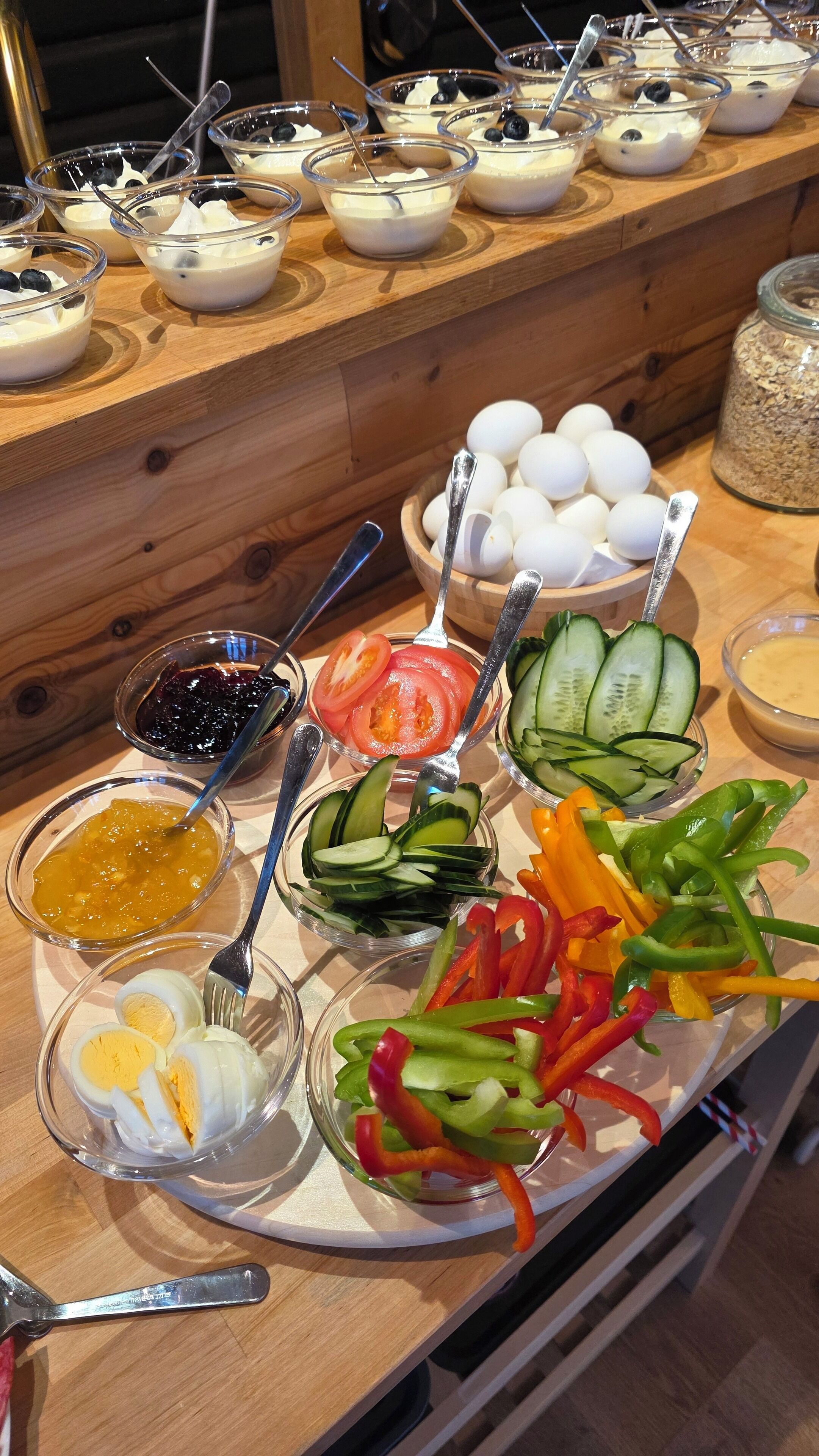 Una mesa de buffet con diversos alimentos, incluyendo tazones de yogur con arándanos, rodajas de pepino, tomates y pimientos, además de un tazón de huevos y un tarro de avena.