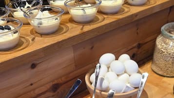 Una mesa de buffet con diversos alimentos, incluyendo tazones de yogur con arándanos, rodajas de pepino, tomates y pimientos, además de un tazón de huevos y un tarro de avena.