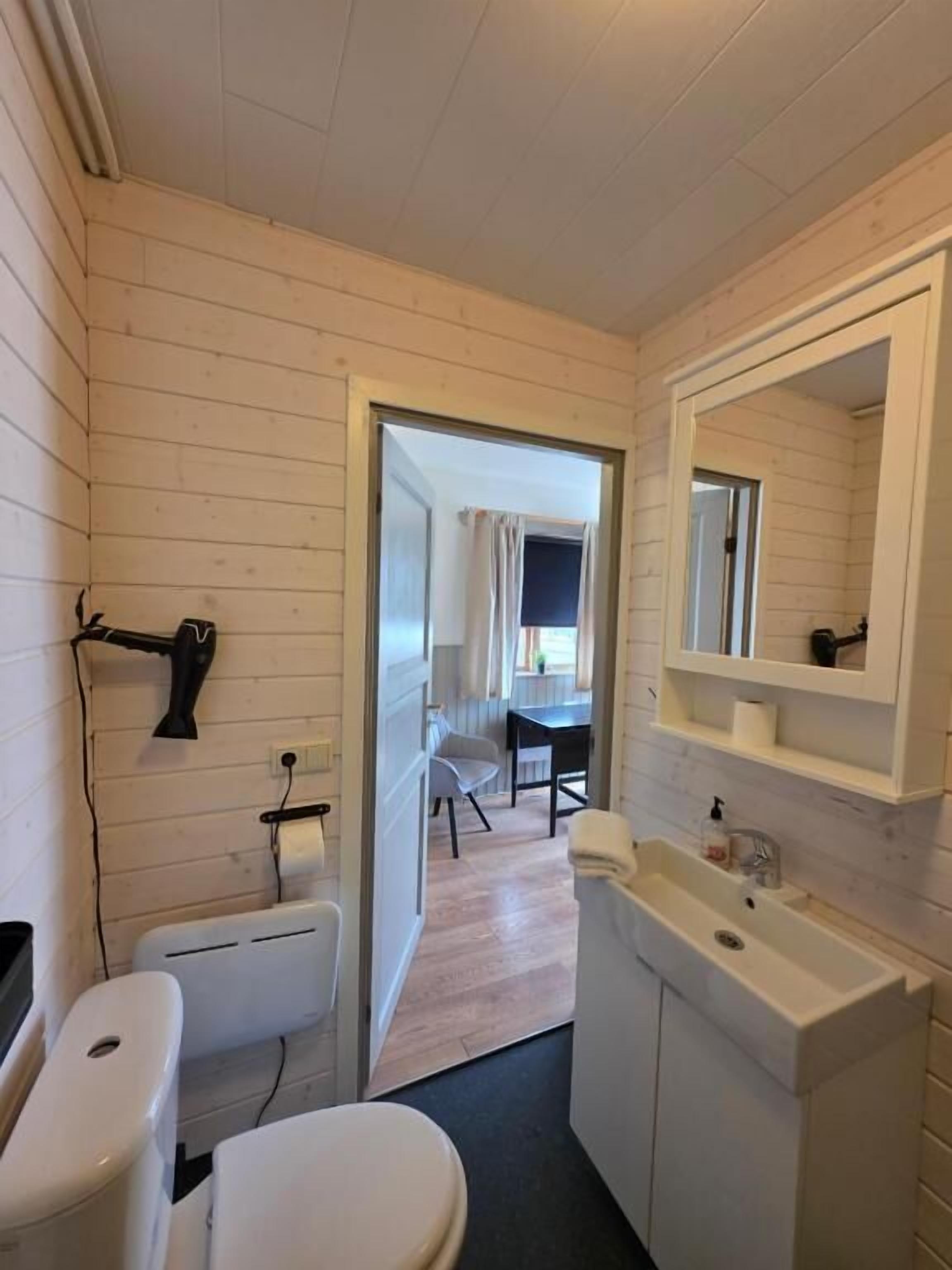 A compact bathroom with a toilet, sink, and mirror. A hairdryer is mounted on the wall, and a door leads to a dining area.