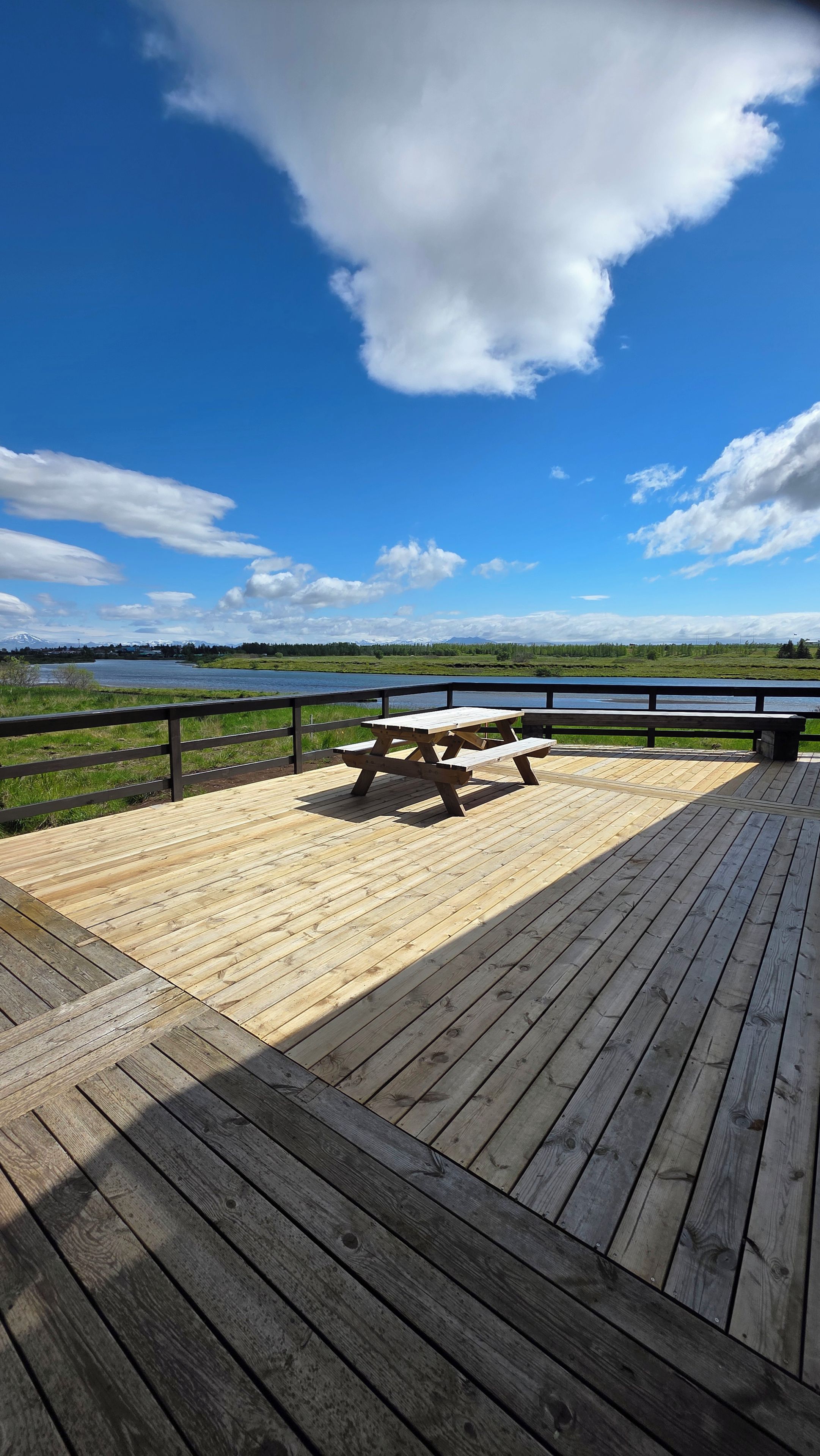 Eine Holzterrasse mit Picknicktisch und Bank, mit Blick auf ein Gewässer und eine grüne Landschaft.