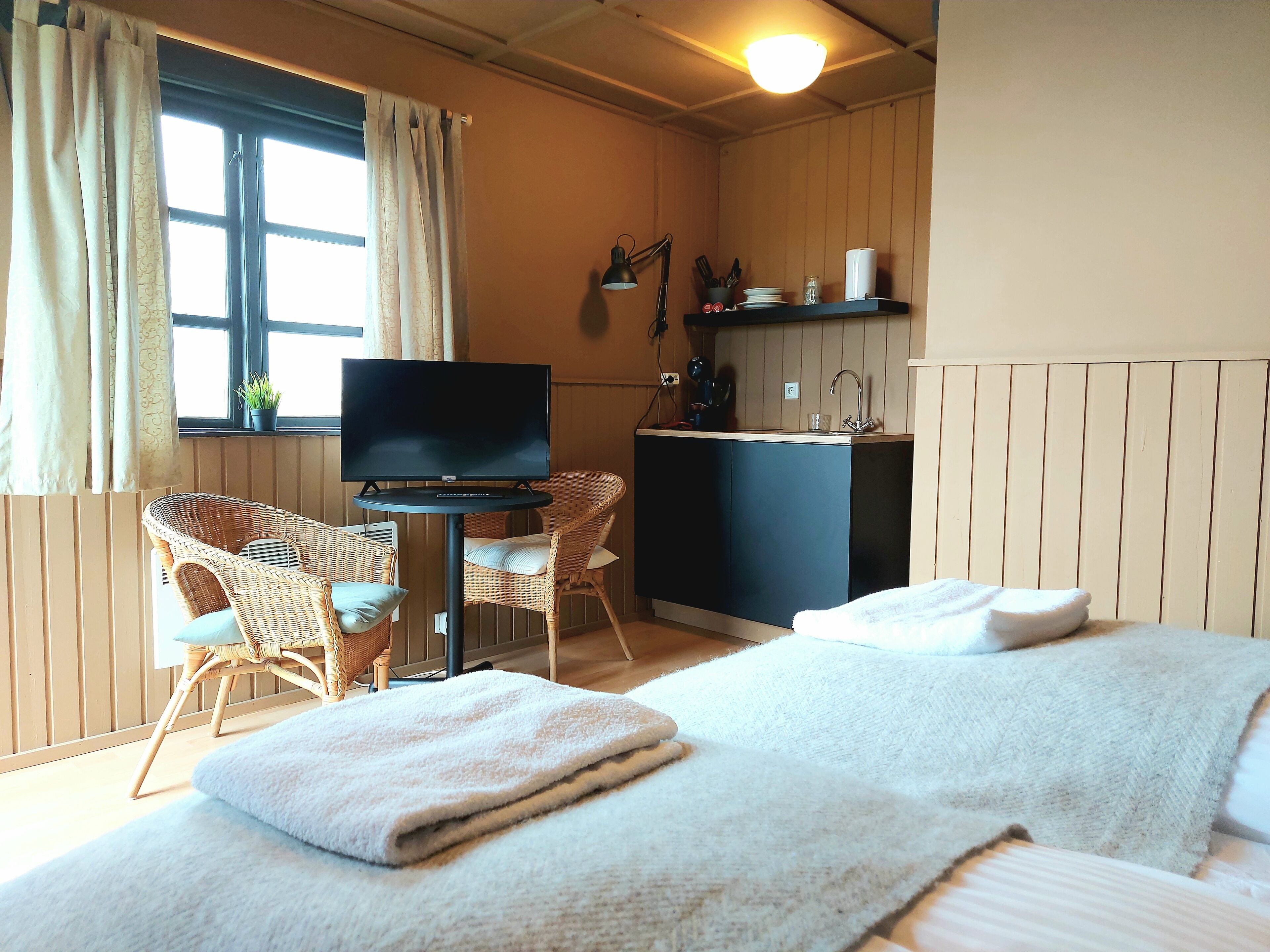 A room with two beds, a television, a small table, wicker chairs, a kitchenette, and a window with curtains.
