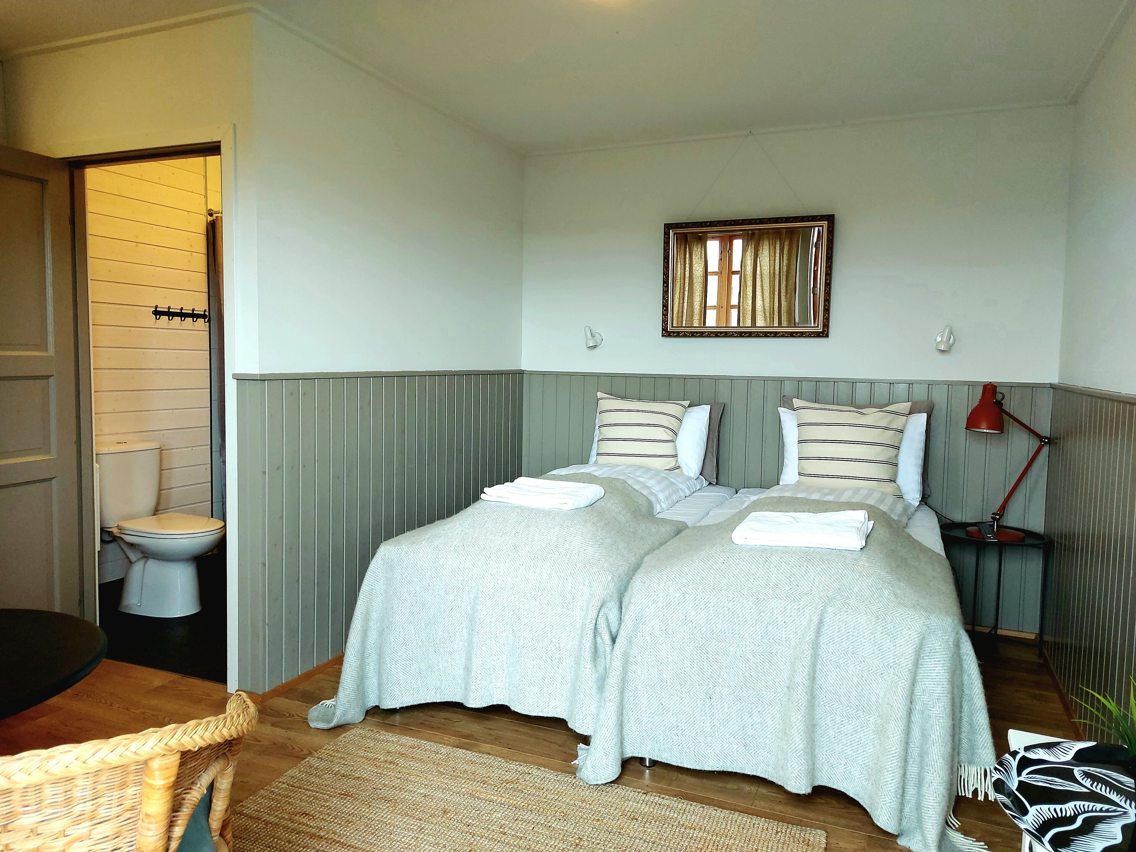 A bedroom with two beds, a bedside table with a lamp, a window with curtains, and a door leading to a bathroom.