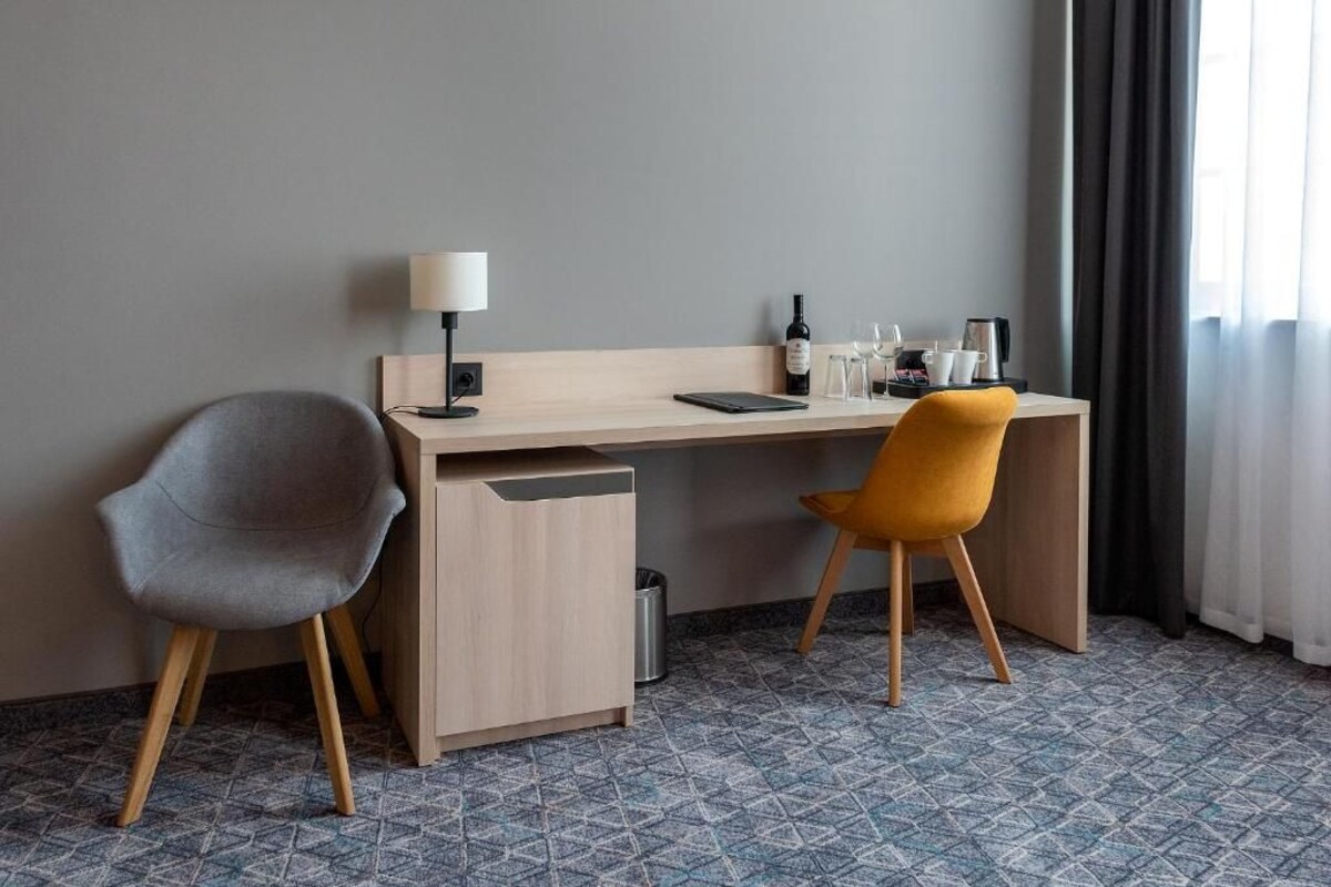 Une chambre d’hôtel équipée d’un bureau, de deux chaises, d’une lampe, d’une bouteille de vin, de verres et d’une poubelle.