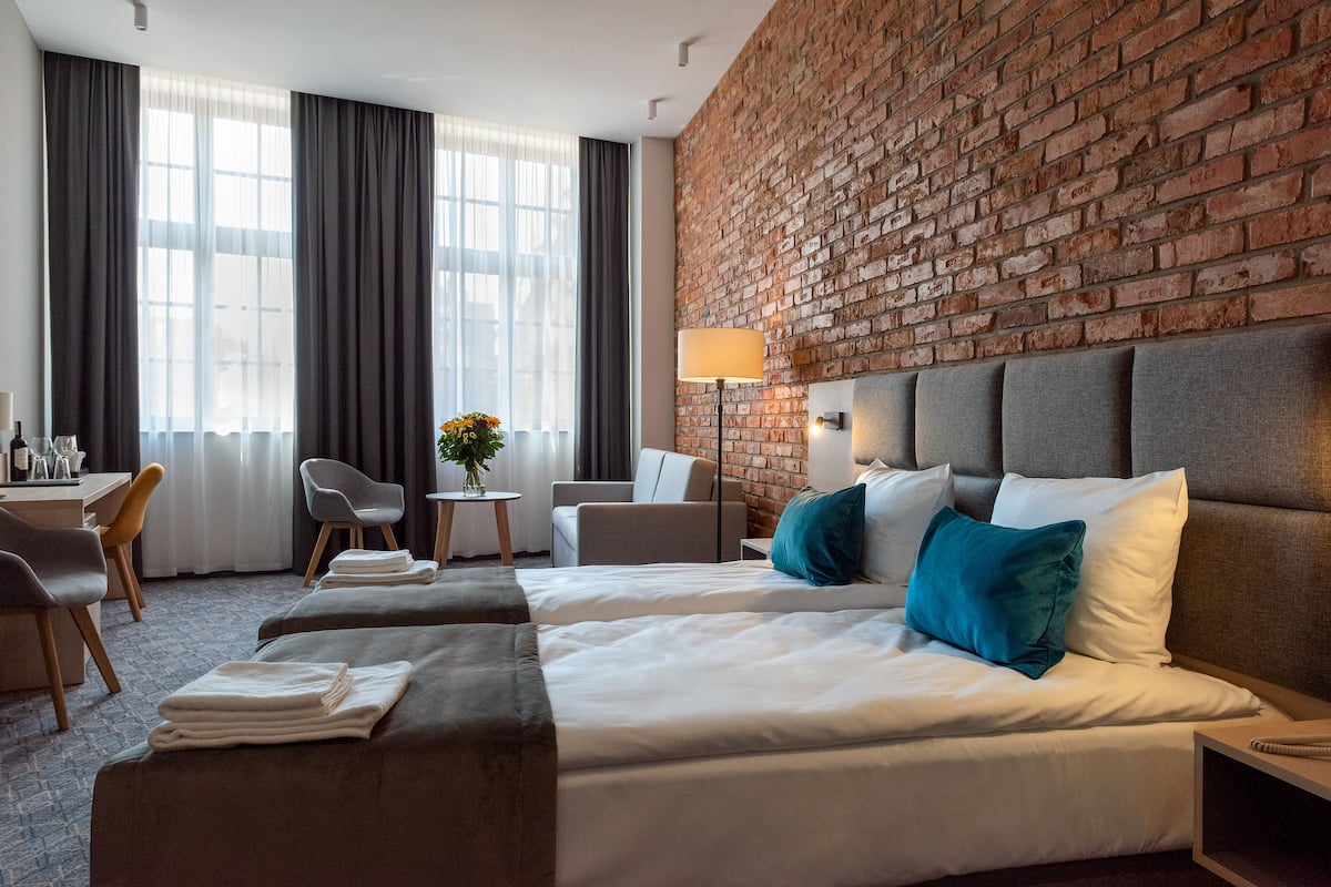Habitación de hotel con dos camas, un escritorio, una silla, una lámpara y una pared de ladrillo.