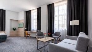 Een moderne hotelkamer met een grijze bank, twee fauteuils, een kleine tafel met vazen, een bureau met stoel, en een bed met een blauw-witte vloerkleden.