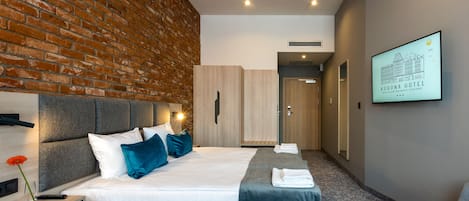 A modern hotel room with a brick wall, a large bed with blue pillows, a wooden wardrobe, and a framed picture on the wall.