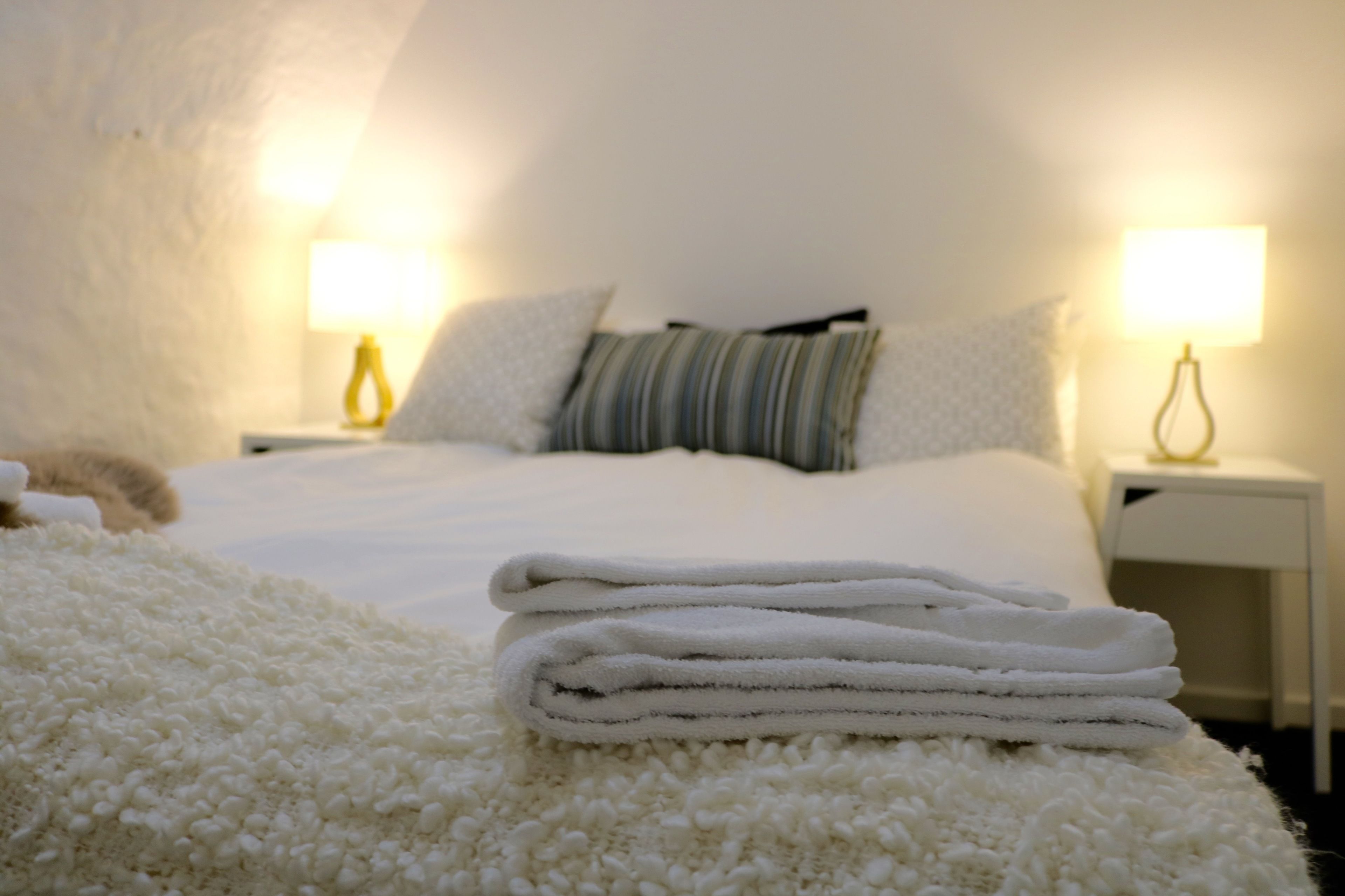 A neatly made bed with white linens and a folded white towel on a fluffy white rug. There are two bedside lamps on white nightstands.
