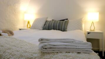 A neatly made bed with white linens and a folded white towel on a fluffy white rug. There are two bedside lamps on white nightstands.