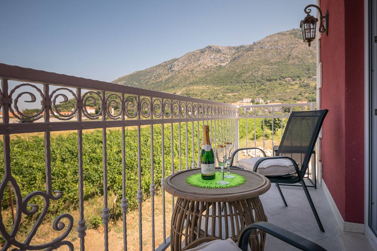 A balcony with a table, chairs, a bottle of wine, and a view of vineyards and hills.