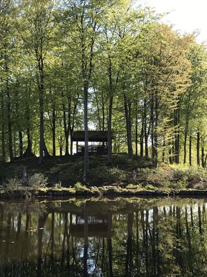 A serene lakeside setting with a wooden structure surrounded by tall trees and a calm water body reflecting the surroundings. - Hillesgården (Klippan)