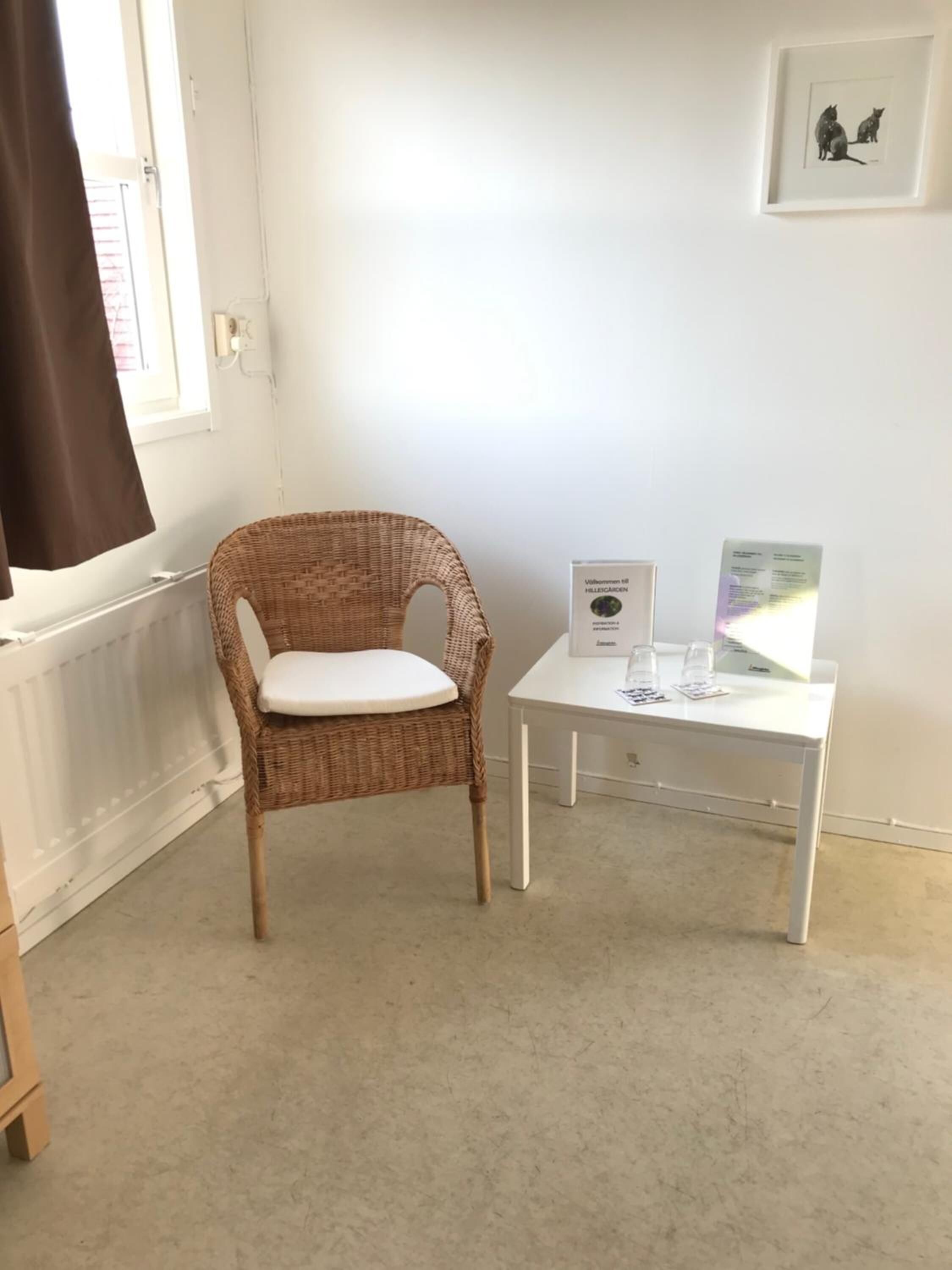 Una silla de mimbre con un cojín blanco en una habitación con un radiador blanco, una mesa blanca con un cuadro enmarcado y una ventana con cortina marrón.