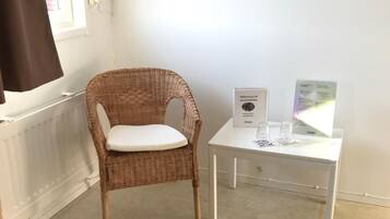 Una silla de mimbre con un cojín blanco en una habitación con un radiador blanco, una mesa blanca con un cuadro enmarcado y una ventana con cortina marrón.