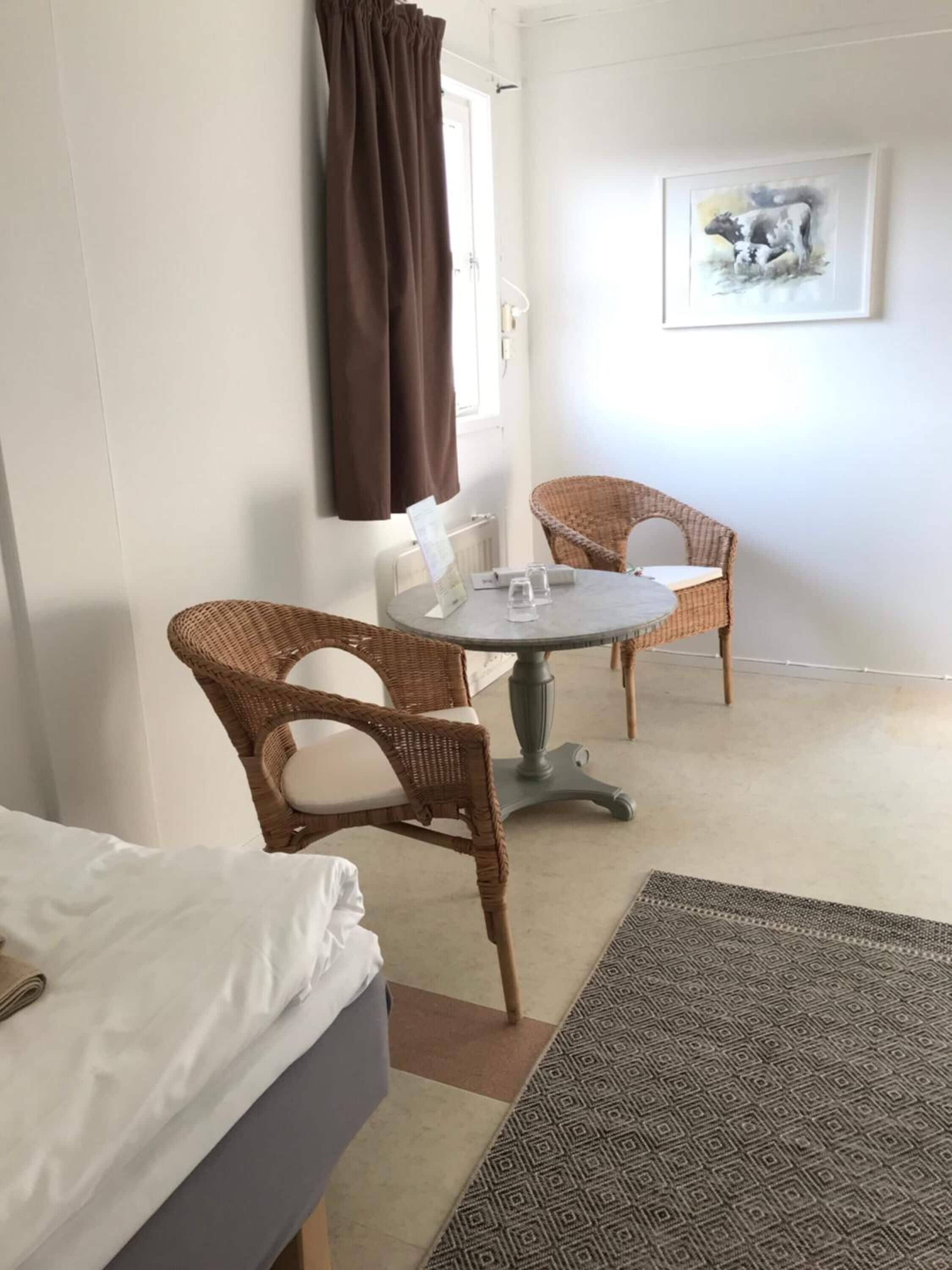 A hotel room with a bed, two wicker chairs, a small round table with a glass, and a framed picture on the wall.