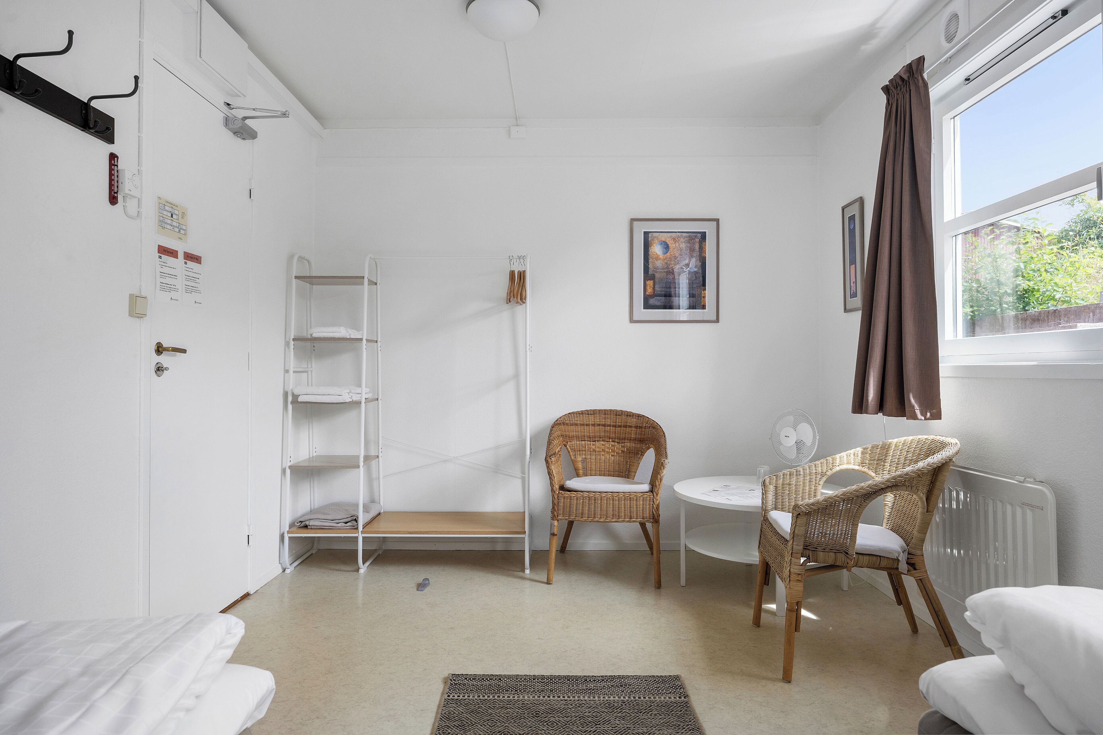 A room with a white bed, a wicker chair, a small table, a radiator, and a window with curtains.