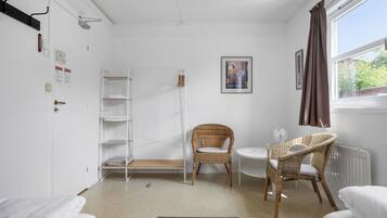 A room with a white bed, a wicker chair, a small table, a radiator, and a window with curtains.