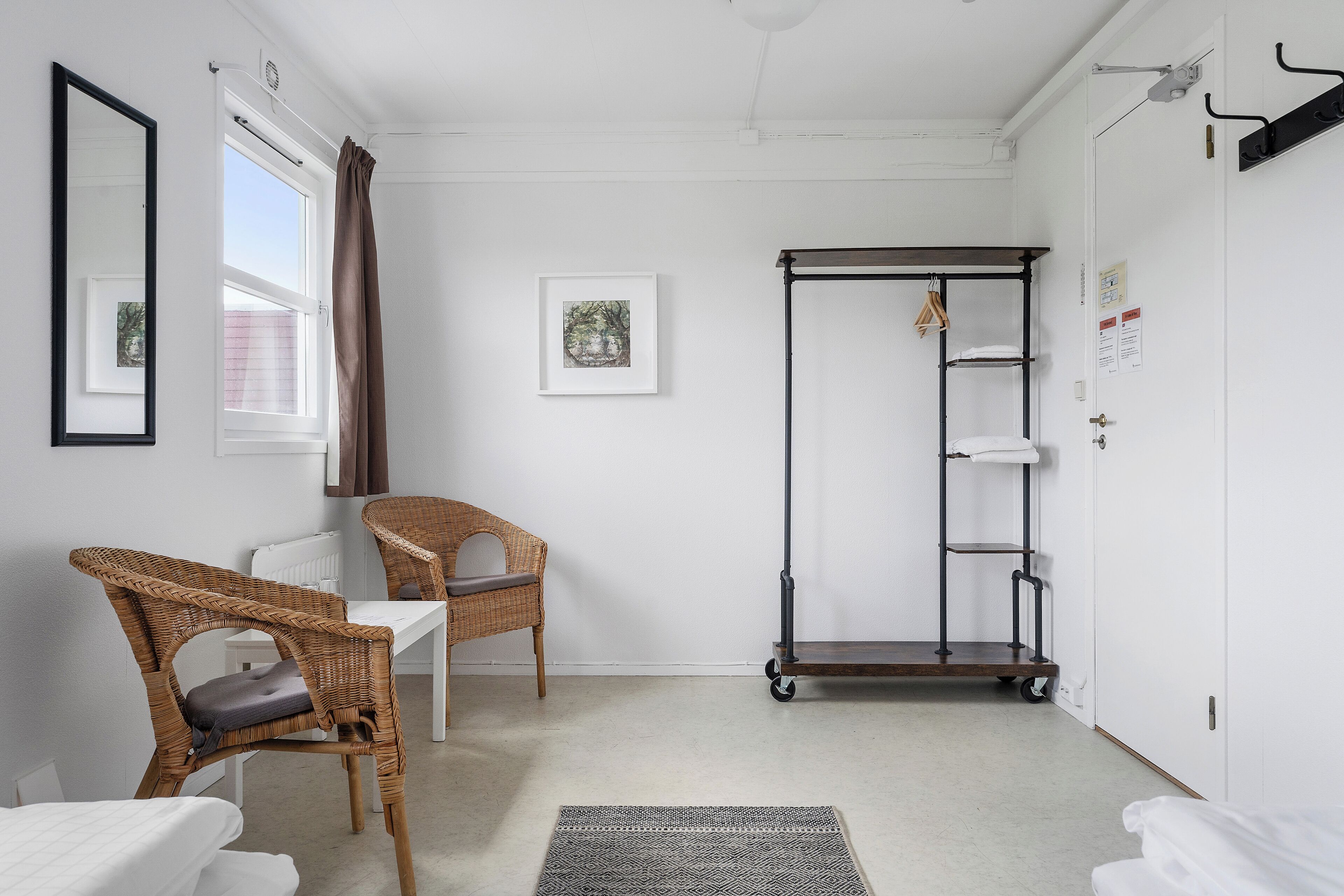 Una habitación con pared blanca, ventana con cortinas, un cuadro enmarcado, una silla de mimbre, una mesita, un perchero metálico y una puerta blanca.