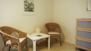 A room with two wicker chairs, a small white table, a bookshelf, and a framed painting on the wall.