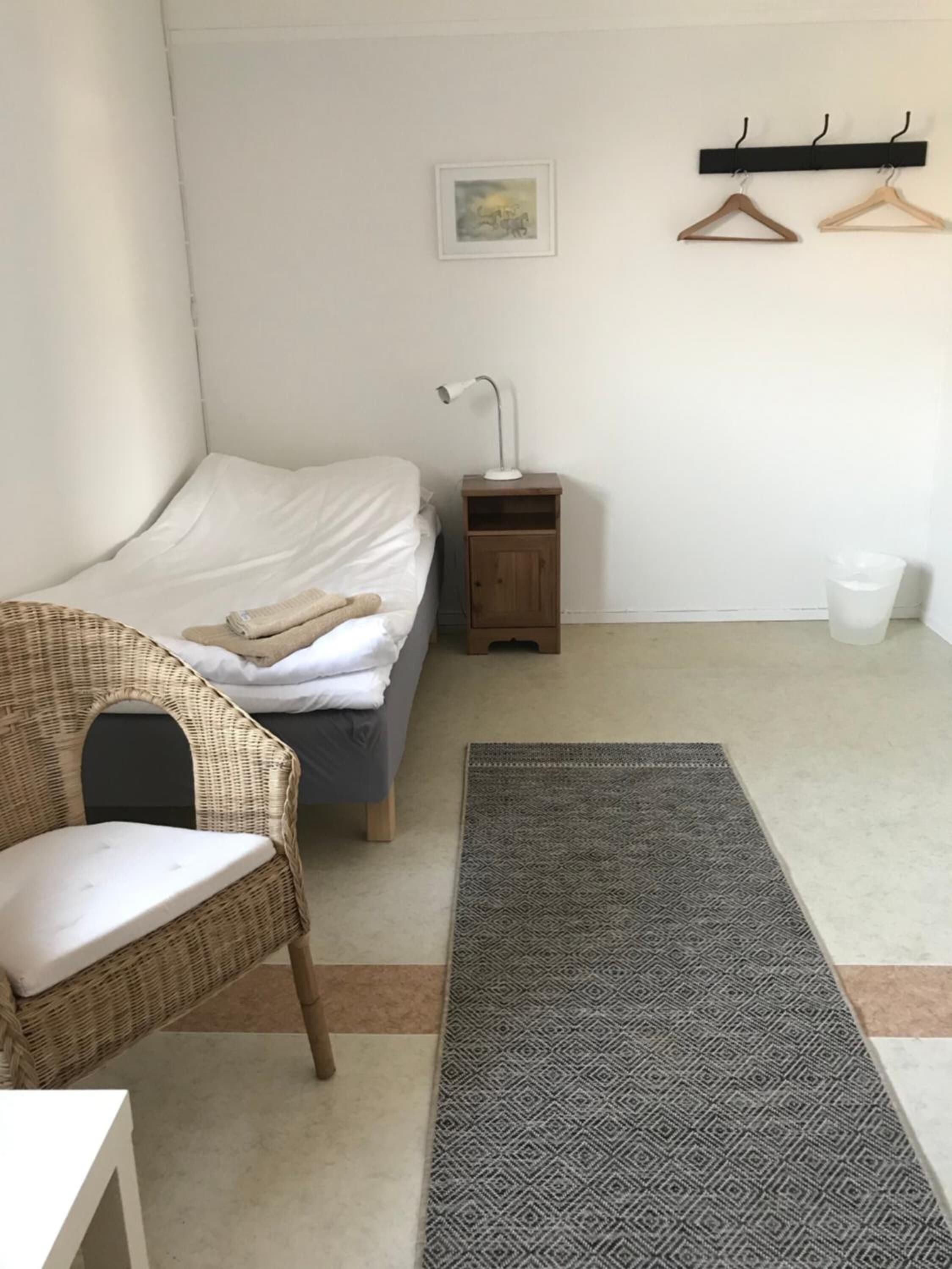 Ein Schlafzimmer mit einem Bett, einem Nachttisch, einem Sessel, einer Pendelleuchte, einem Teppich und einem Kleiderhaken.