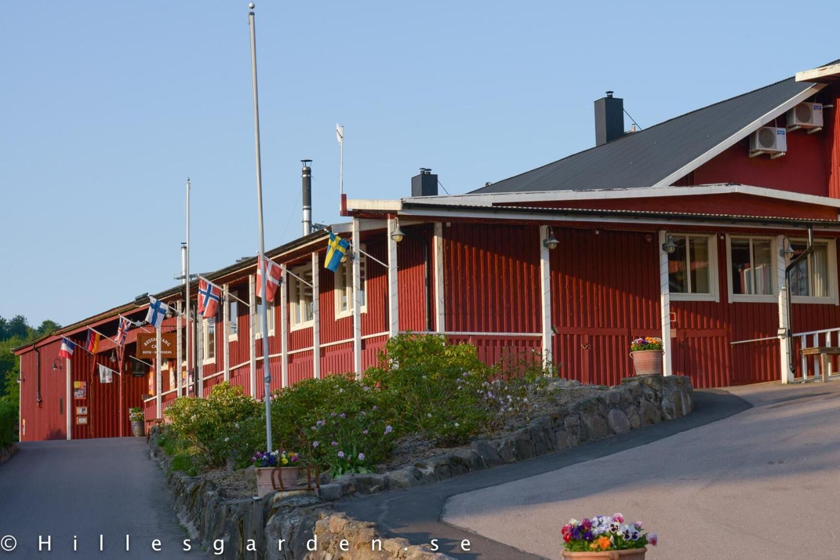 A red building with a sloped roof, multiple flags, and a stone wall with flowers.