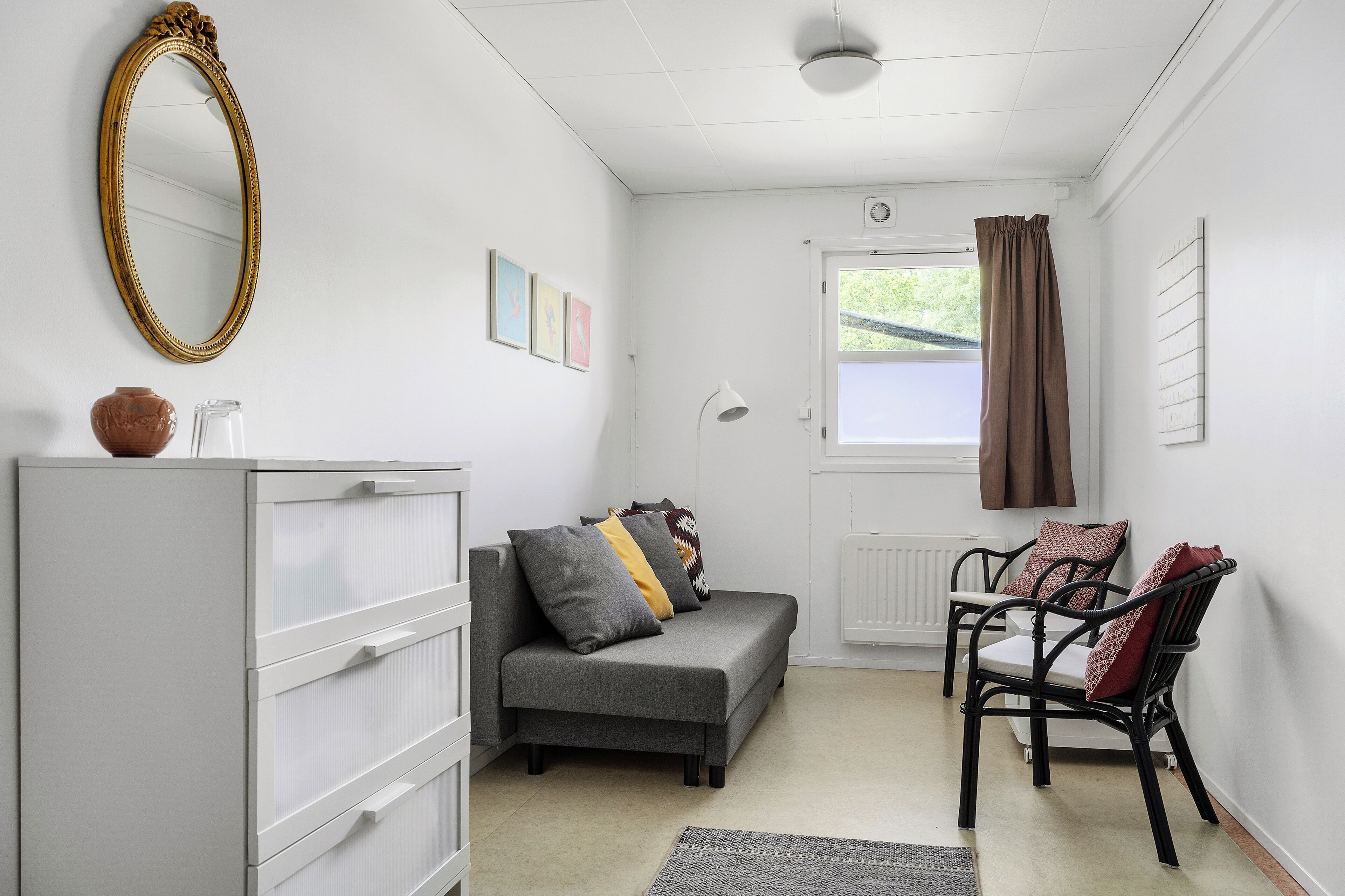 A room with a grey sofa, a white cabinet, a black chair, a mirror, and a window with curtains.