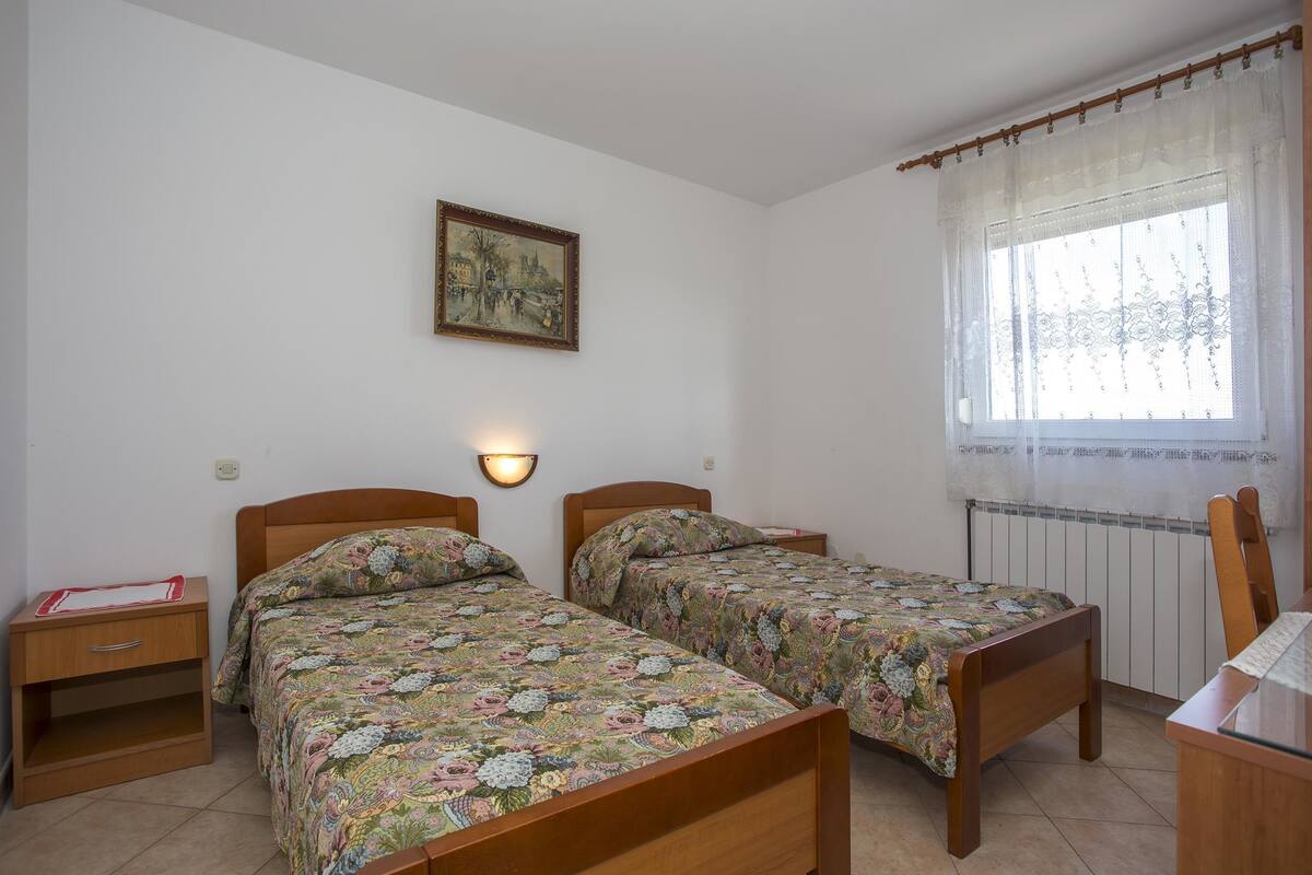 Una habitación con dos camas individuales, una mesita de noche con lámpara, un cuadro enmarcado en la pared y una ventana con cortinas traslúcidas.