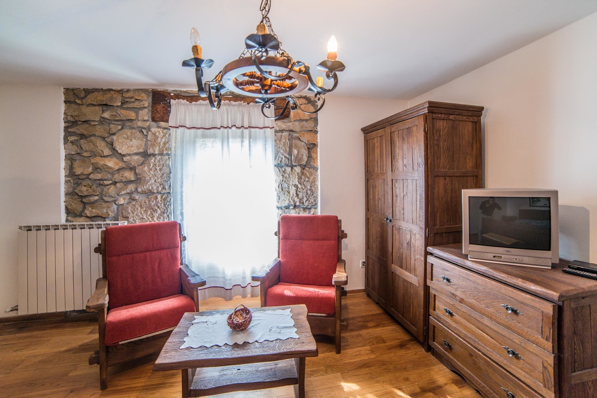 A living room with two red armchairs, a wooden coffee table, a stone wall, a wooden cabinet, and a television.