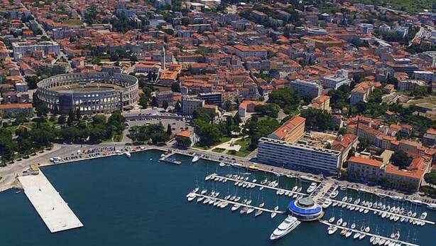 Vista aérea de una ciudad costera con un edificio redondo destacado, probablemente un sitio histórico, y un puerto deportivo con numerosas embarcaciones amarradas.