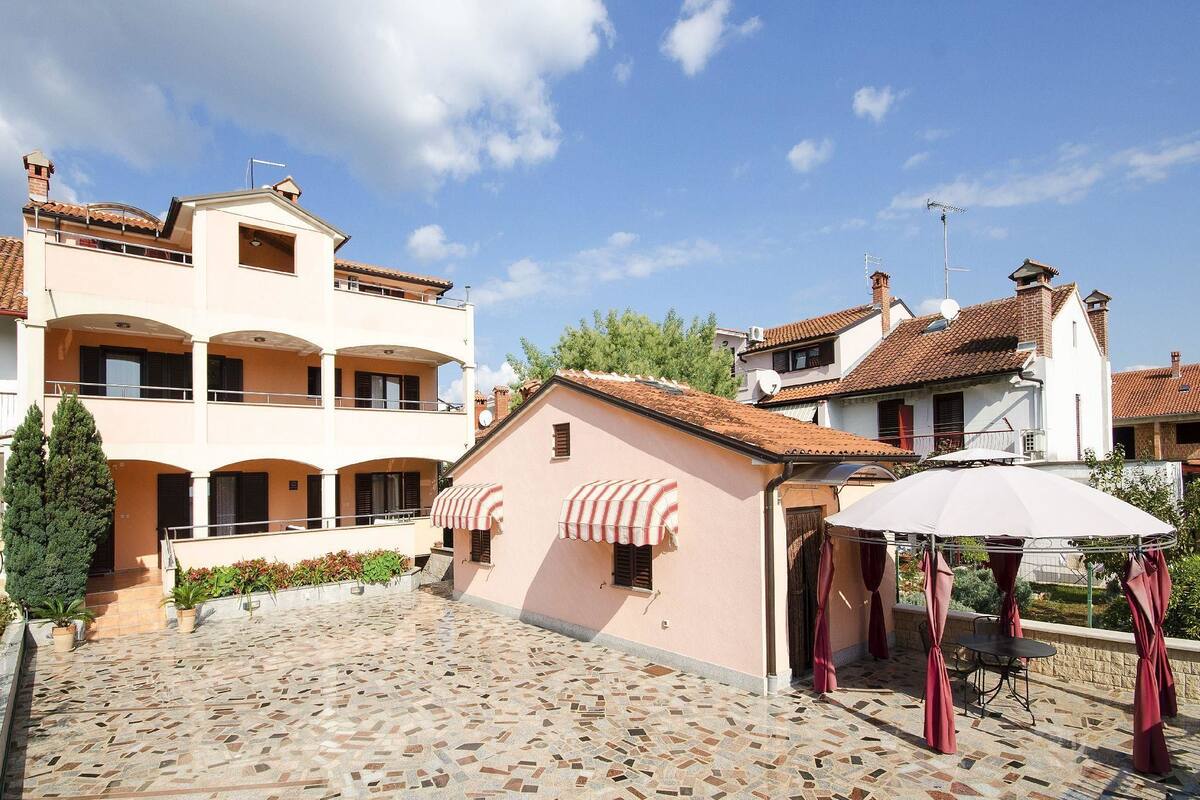 A courtyard with a tiled floor, a building with a balcony, and a small structure with a roof.