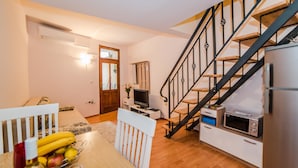 A compact living space with a wooden dining table, a TV, a microwave, and a staircase with black railings.