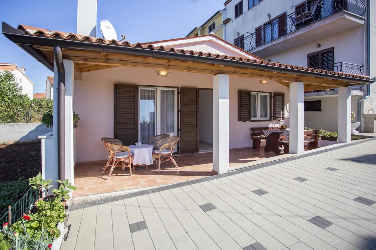 A covered outdoor patio with wicker furniture and a table, adjacent to a building with a tiled roof and balconies.