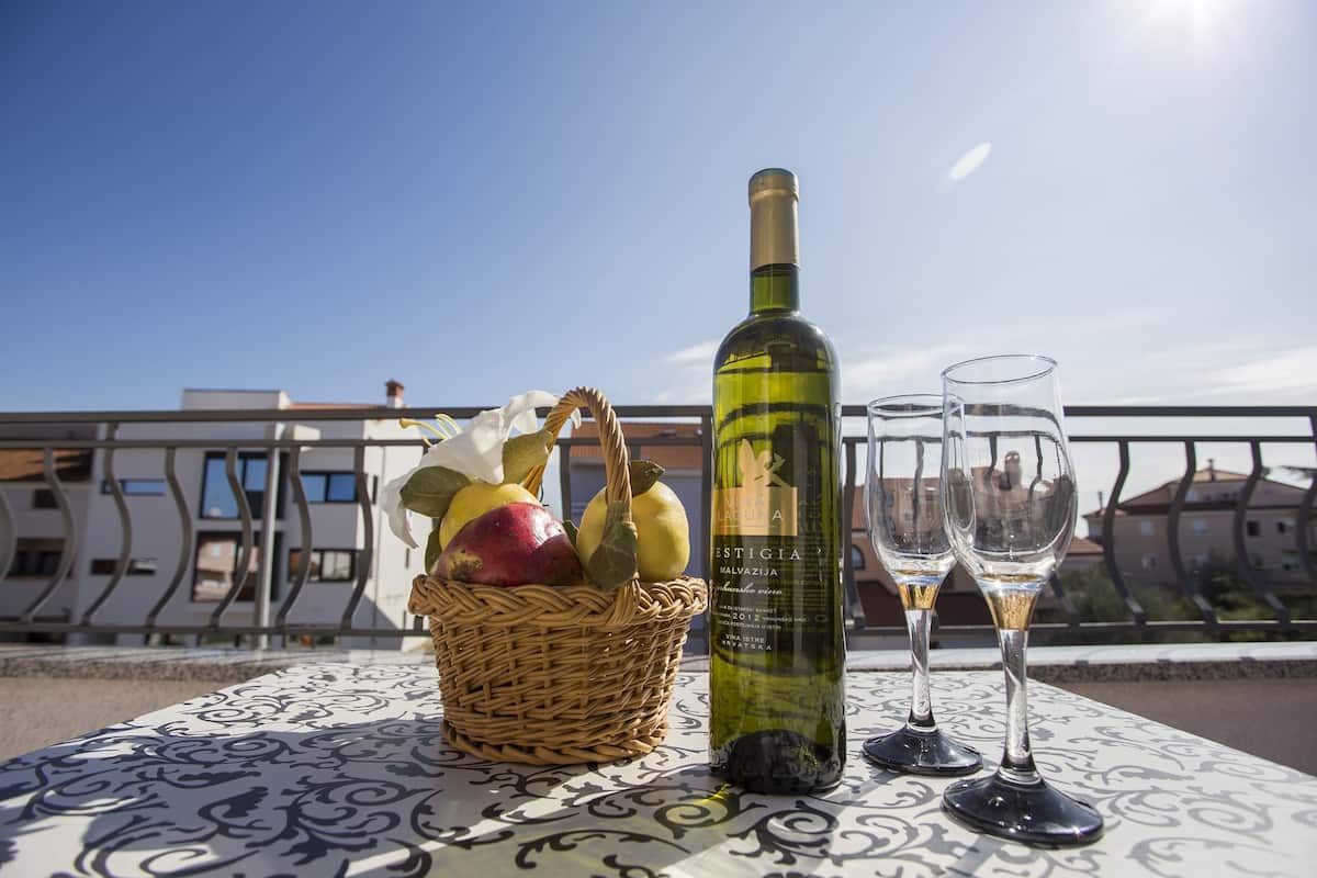 A table with a bottle of wine, two glasses, and a basket of fruit on a balcony with a railing and buildings in the background.