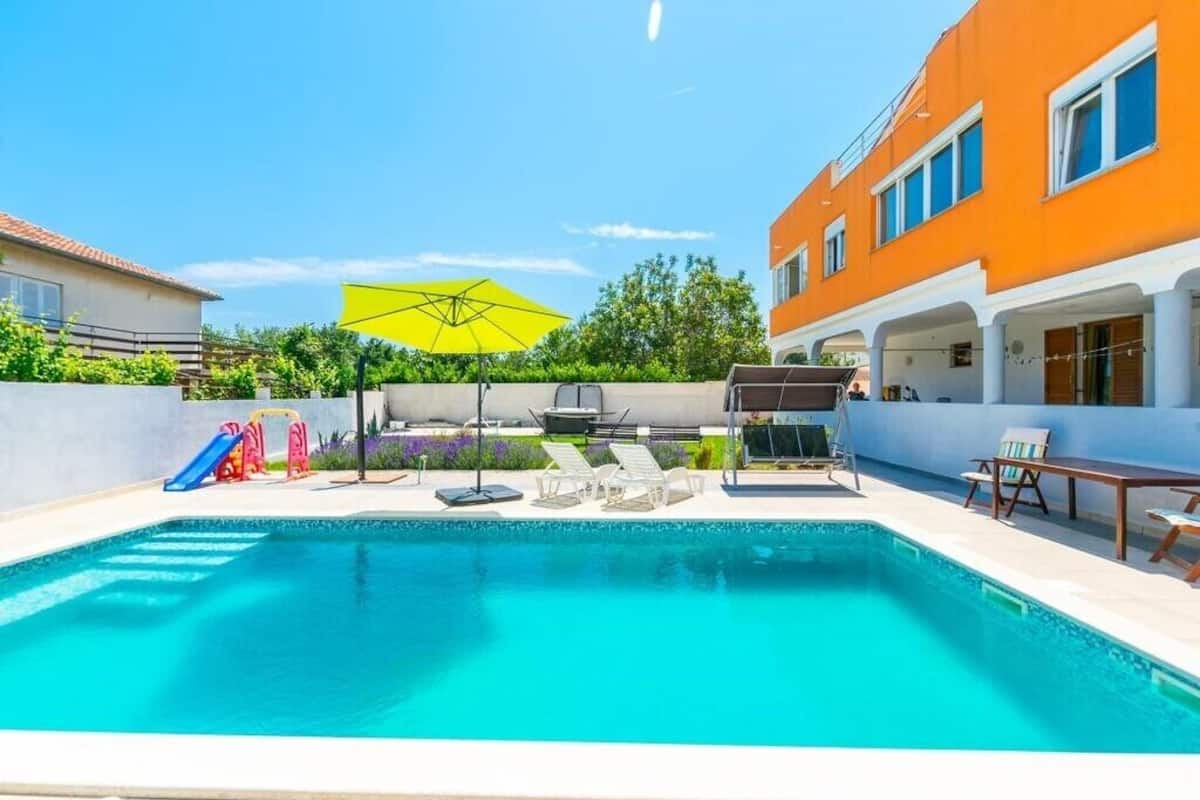 A swimming pool with lounge chairs and a slide, surrounded by a white wall and an orange building.