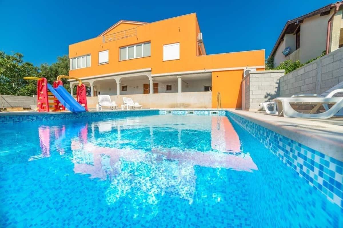 A swimming pool with a slide, a covered patio area, and a two-story building with an orange facade.