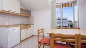 A compact kitchen with white cabinets, a sink, and a refrigerator. There is a dining table with chairs and a window with a view of buildings. - Apartment Vinko (Sibenik)