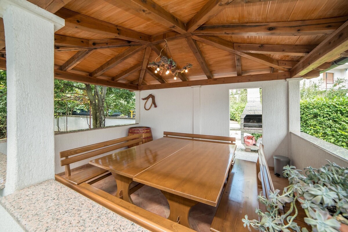 Un espace extérieur abrité, aménagé pour les repas, avec une table et des bancs en bois, un luminaire suspendu et une vue sur un barbecue situé à l’extérieur.