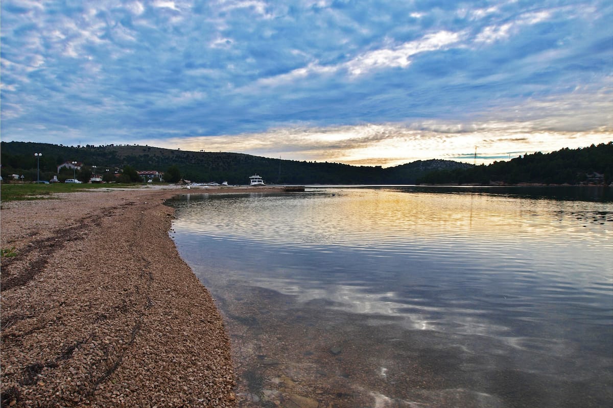 A pebbled beach with calm water, a boat, and a distant hill with buildings.