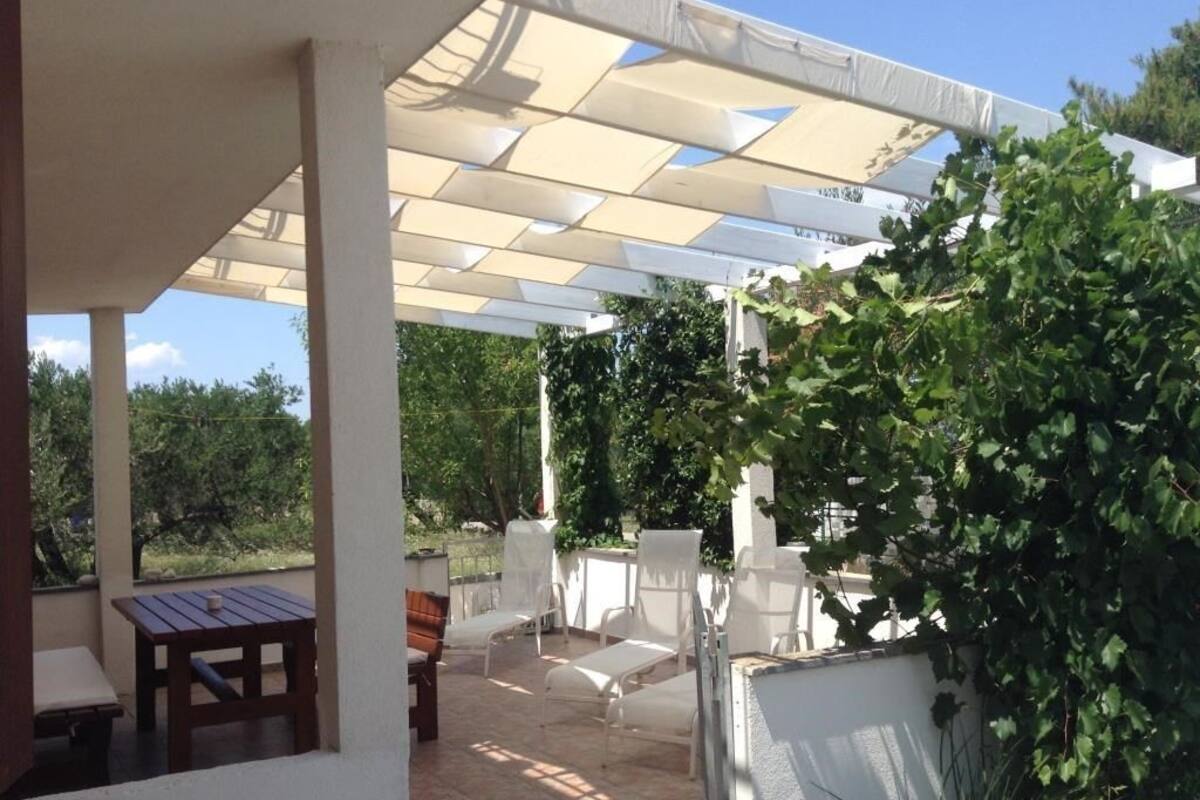 A covered outdoor seating area with a table and chairs, surrounded by greenery.