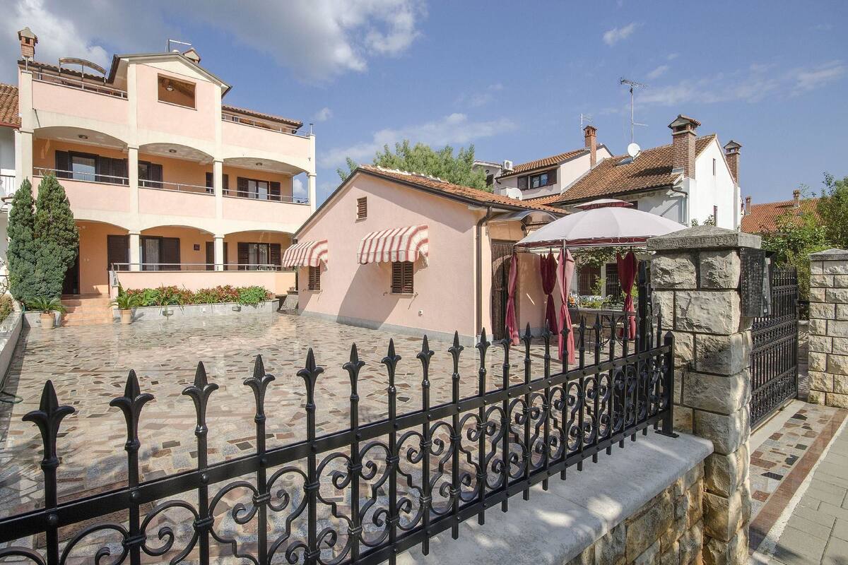 A courtyard with a stone fence, a small building with a red awning, and a multi-story building with balconies.