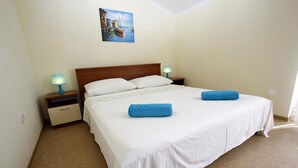 A neatly made bed with white linens and two blue towels, a wooden headboard, and a painting of a coastal scene above it. - Apartments Vesna Mimice (Omis)