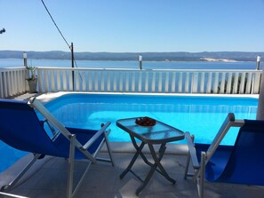 A swimming pool with a table and chairs, surrounded by a white fence, with a view of the sea and distant hills. - Apartments Vesna Mimice (Omis)