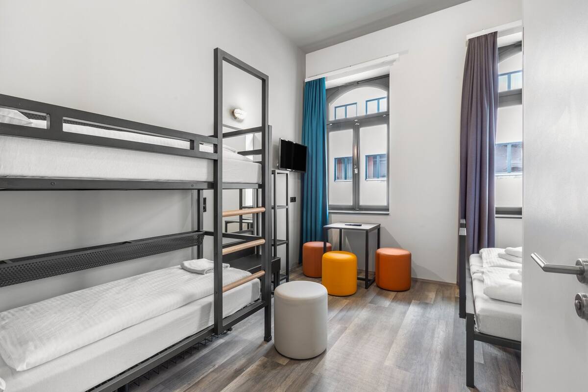A modern hotel room with bunk beds, a television, a small table, and orange ottomans.