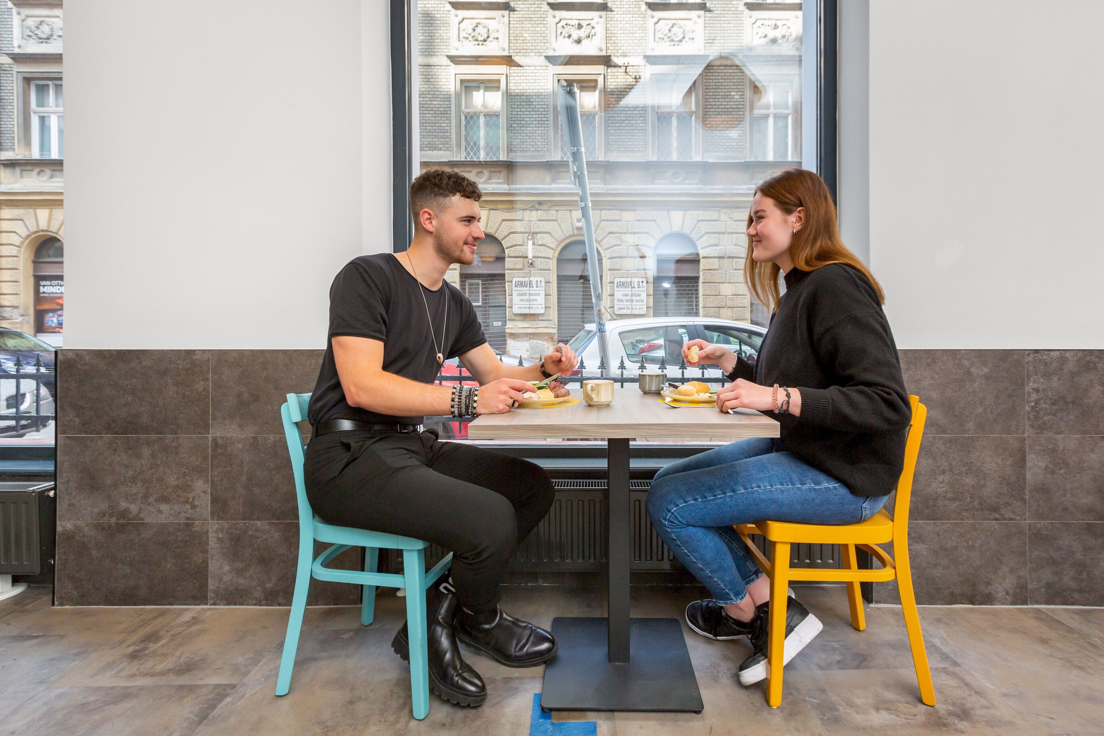 Two people sitting at a table in a cafe with a view of a building through the window.