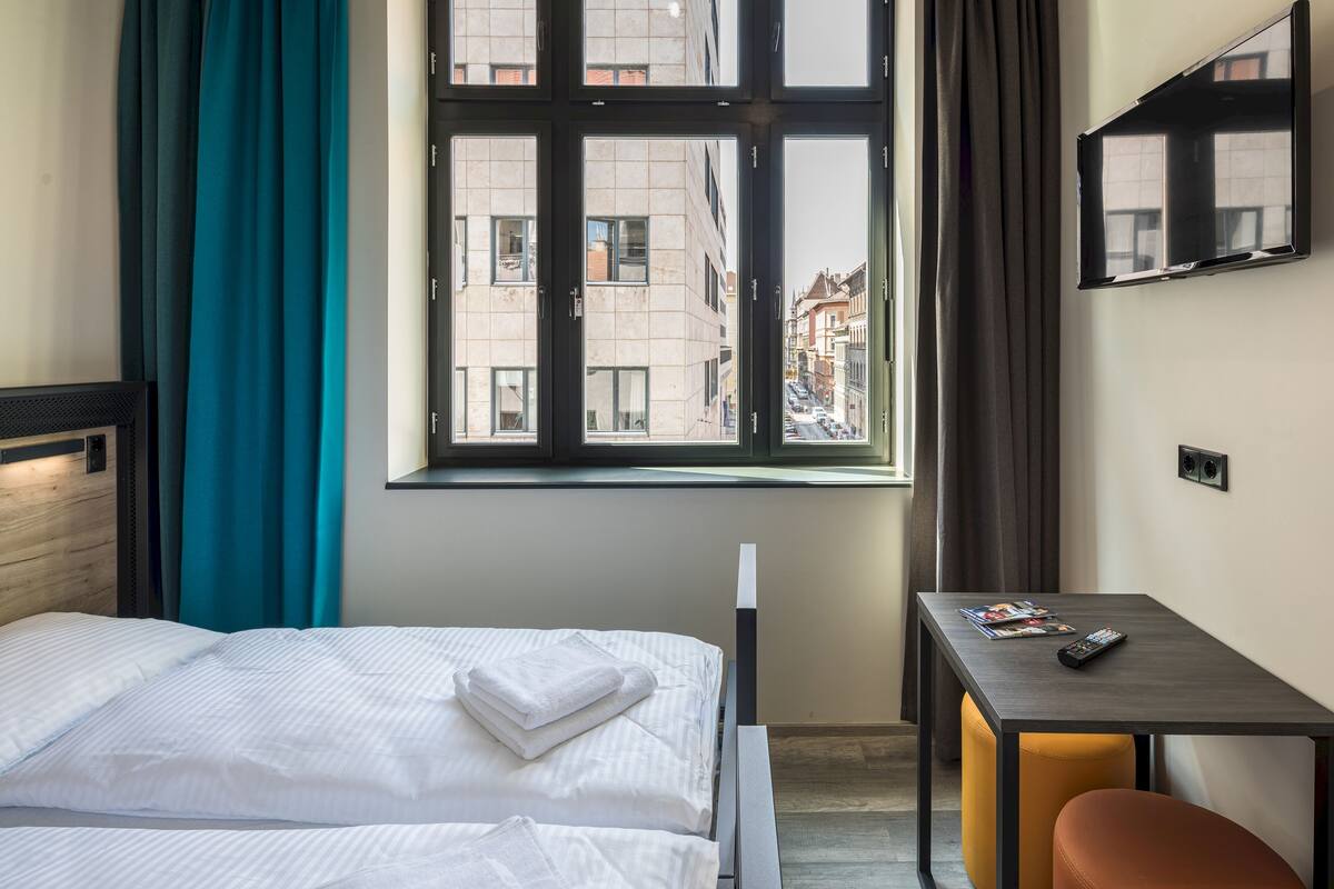 A hotel room with a bed, a desk, a chair, and a window with a view of buildings.