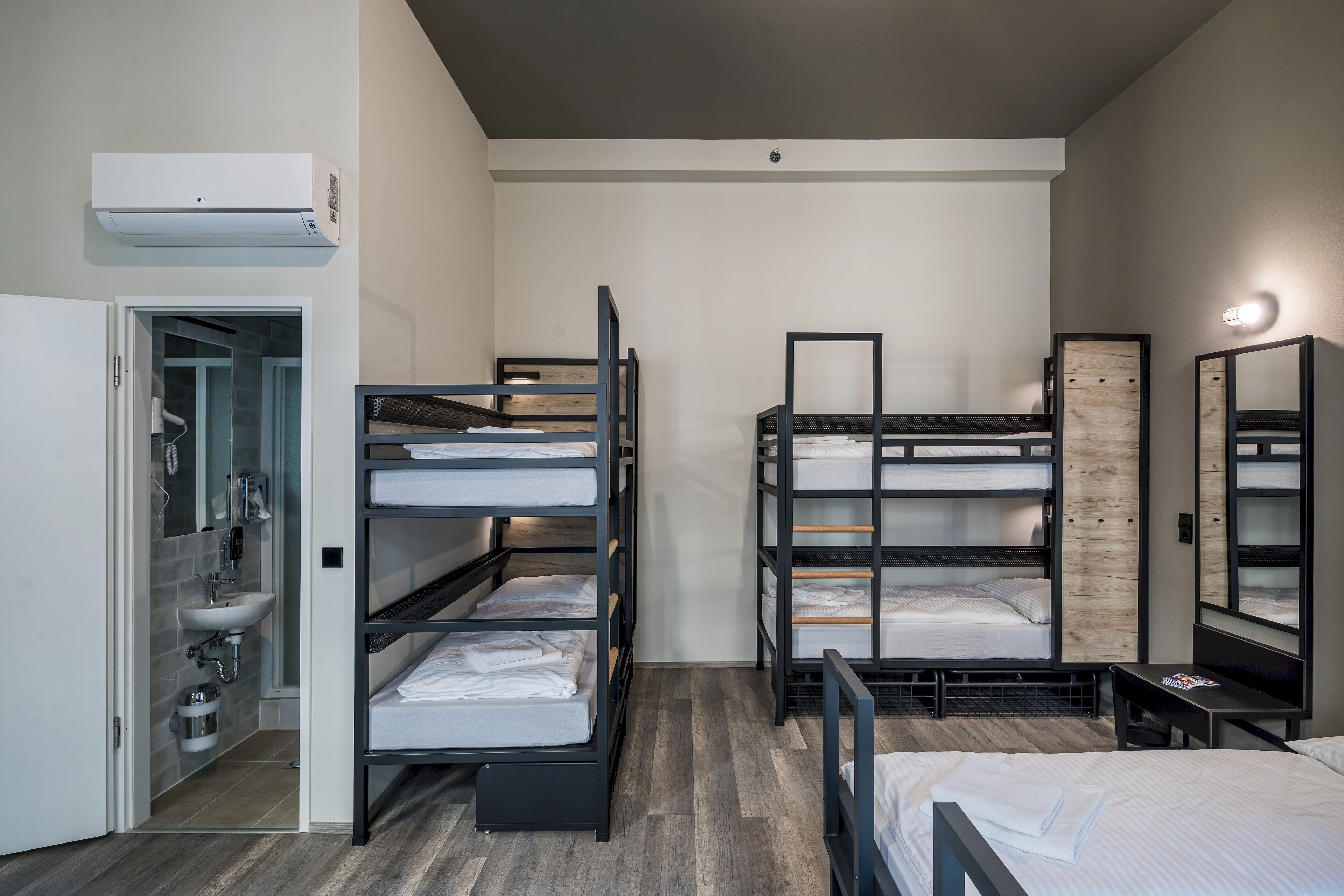 A modern hotel room with bunk beds, a small desk, a mirror, and a bathroom with a sink and shower.