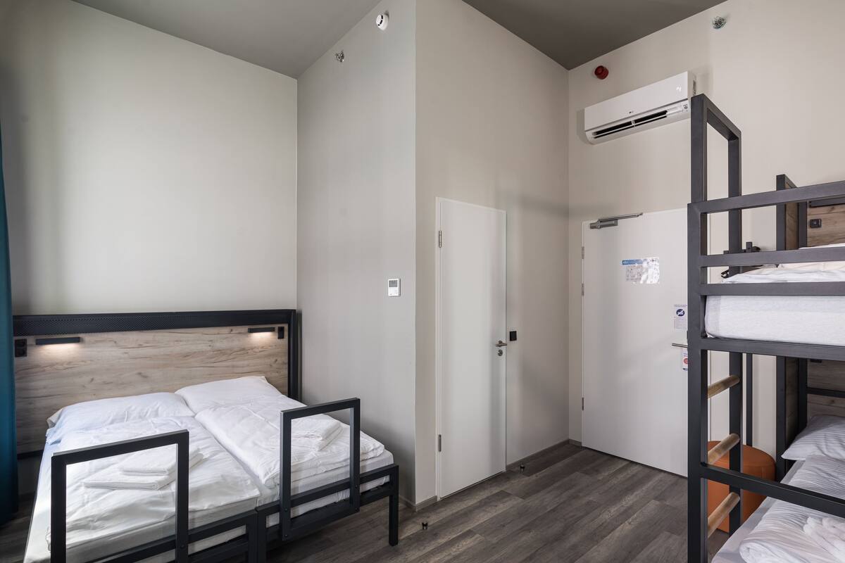 A compact hotel room with a bunk bed, a single bed, a wardrobe, and a small seating area.