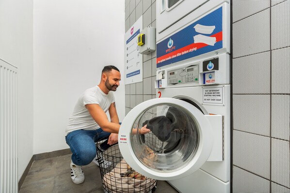 A person crouching next to a front-loading washing machine with a basket of clothes inside.