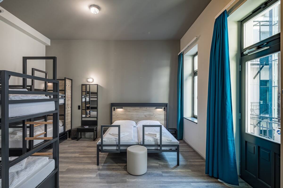 A compact hotel room with a bunk bed, a single bed, a bedside table, a mirror, and a window with curtains.